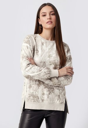 Junge Frau mit langen glatten Haaren trägt ein helles Sweatshirt mit Blumenmuster und schwarze Lederhose, Arme verschränkt, neutraler Hintergrund.