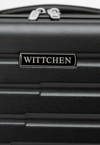 Černý tvrdý kufr s vertikálními drážkami, se stříbrným zipem a černou jmenovkou s vyraženým nápisem "WITTCHEN."
