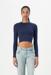 Navy long-sleeve crop top with a round neckline and embossed logo. Worn with light blue high-waisted denim jeans. Smooth fabric texture.
