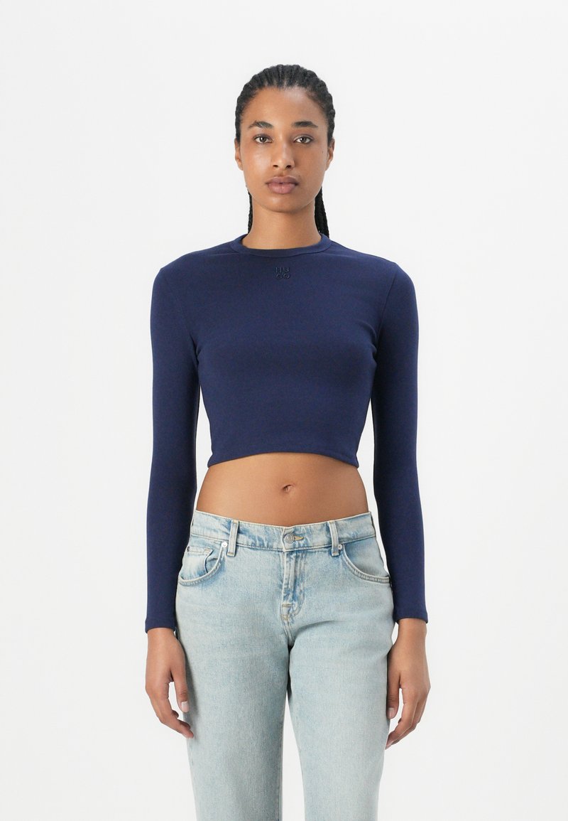 Navy long-sleeve crop top with a round neckline and embossed logo. Worn with light blue high-waisted denim jeans. Smooth fabric texture.
