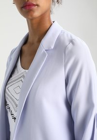 Veste blazer lavande claire avec col cranté, en tissu lisse. Présente une coupe cintrée et des manches légèrement bouffantes, portée sur un haut à motif.