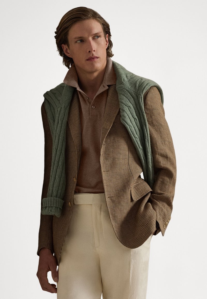 Braun karierter Blazer über einem beigen Poloshirt, dazu ein grüner, grob gestrickter Pullover, der über die Schultern gelegt ist. Cremefarbene Hosen runden den Look ab.