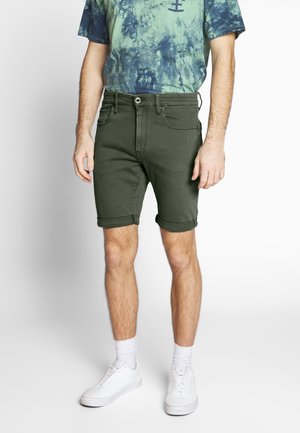 Homme portant un short vert foncé retroussé, un t-shirt tie-dye bleu, des chaussettes blanches et des baskets blanches, debout sur un fond clair.