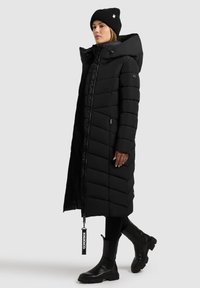 Lang zwarte pufferjas met een chevronpatroon, brede kap, rits aan de voorkant en zijzakken. Draagt een bijpassende gebreide beanie en zwarte laarzen.