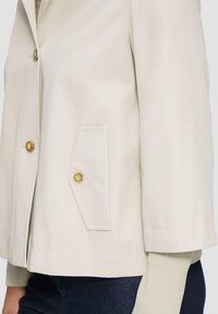 Beige jack met een knoopsluiting, twee voorkruiszakken en een geribbelde mouwdetail. Gemaakt van een gladde stof met een gestructureerd ontwerp.