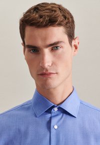 Jasnoniebieska koszula z długim rękawem, z kołnierzem rozpostartym, o teksturowanej tkaninie i białych guzikach. Zbliżenie na górną część torsu męskiego modela.