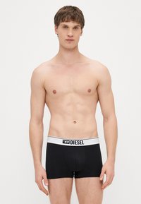 Melni boksa šorti ar baltu elastīgu vidukļa joslu, kurā redzams "DIESEL" logotips; cieši pieguļošs dizains, gluda auduma tekstūra un klasiskā griezuma.