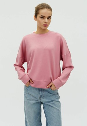 Mlada ženska s svetlo rjavimi lasmi, spetimi v figo, oblečena v ohlapno roza majico in svetlo modre kavbojke, z rokami v žepih, nevtralen ozadje.