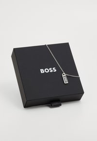 Ασημένιο κολιέ με ορθογώνια ετικέτα σε λεπτή αλυσίδα, εκτιθέμενο σε μαύρο κουτί με το όνομα της μάρκας "BOSS" σε λευκό.