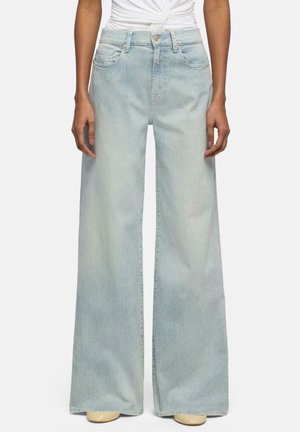 Frau trägt hellblaue Jeans mit Nadelstreifen und weitem Bein sowie ein weißes, gerafftes ärmelloses Oberteil, steht mit entspannt an den Seiten hängenden Händen.