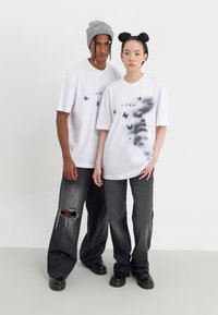 Camisetas oversize blancas con estampados de mariposas y la palabra "VISIÓN" en negro, combinadas con jeans oscuros desgastados y zapatos negros.