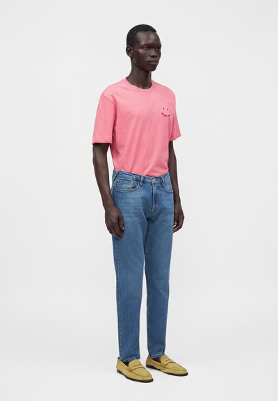 Homme debout portant un t-shirt rose à manches courtes, un jean bleu et des mocassins jaunes sur un fond clair uni.