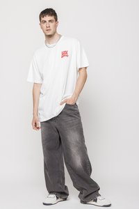 T-shirt bianco oversize con testo rosso, abbinato a jeans grigi larghi. Sneakers bianche con dettagli blu navy. Design semplice, moderno e informale.