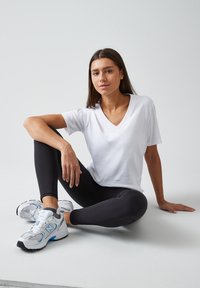 Weißes V-Ausschnitt-T-Shirt kombiniert mit schwarzen Leggings und grauen Sportschuhen mit blauen Akzenten. Der Stoff wirkt glatt und figurbetont.