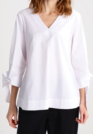 Kobieta ubrana w białą bluzkę z dekoltem w kształcie litery V i rękawami 3/4 ozdobionymi związanymi kokardami, w połączeniu z czarnymi spodniami na jednolitym tle.