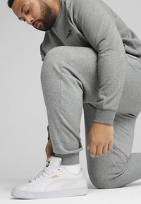 Grå sweatshirt og sweatpants, tekstureret stof. Hvide Puma sneakers med gule accenter. I knæposition, med fokus på at snøre skoene.