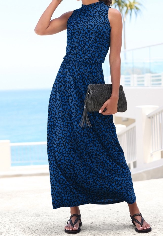 Femme portant une robe maxi bleue à motifs sans manches, tenant une pochette noire, debout à l'extérieur près d'une rambarde en bord de mer.