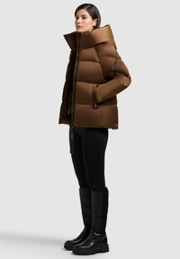 Giacca imbottita marrone con colletto alto, dotata di zip frontale e tasche laterali. Modello abbinato a stivali neri sopra il ginocchio e pantaloni.