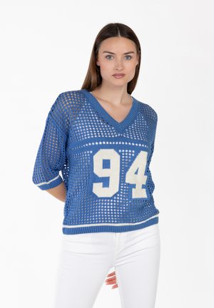 Kobieta w niebieskim siateczkowym swetrze z białą liczbą 94 i białych spodniach, stojąca na tle jednolitej białej ściany.