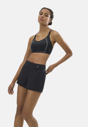 Mujer con sujetador deportivo negro y shorts deportivos de cintura alta, apoyada contra un fondo claro y liso, con una mano en la espalda.