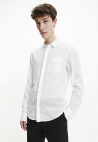 Camicia button-up bianca in lino con taschino sul petto, maniche lunghe e orlo arrotondato, esposta su uno sfondo neutro.