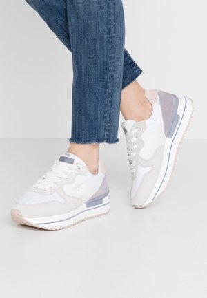 Voeten die witte en grijze sneakers met veters dragen, gecombineerd met blauwe cropped jeans tegen een effen grijze achtergrond.