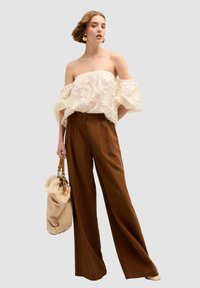 Top bianco scollato con texture floreale, abbinato a pantaloni larghi marroni; tiene in mano una borsa beige con un accento in pelliccia sintetica.