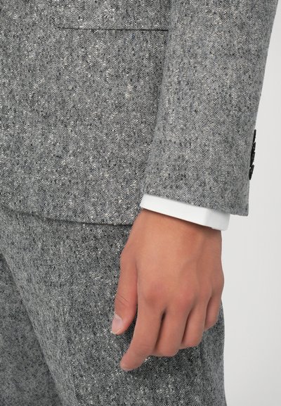 Grau meliertes Anzuggewebe mit strukturierter Oberfläche, wobei ein weißer Hemdärmel unter dem Jackenärmel hervorschaut. Schwarze Knopfdetails sind sichtbar.