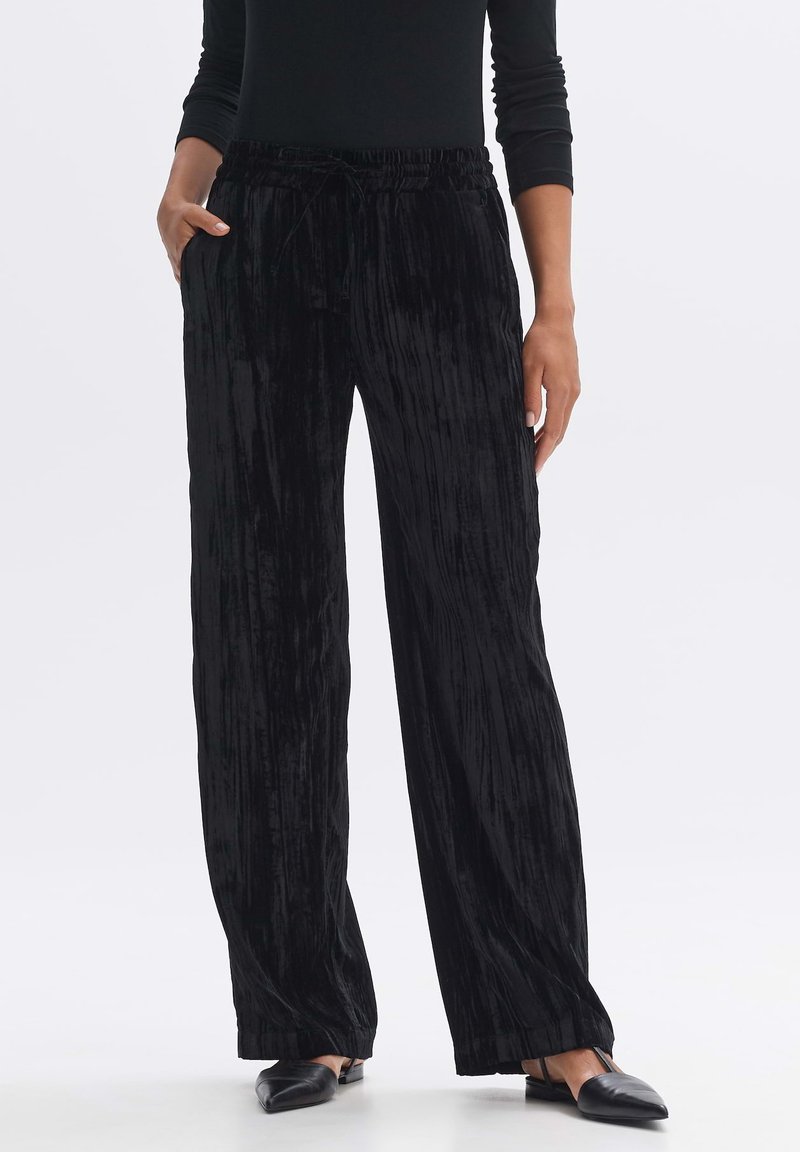 Black velvet trousers with a pleated texture, featuring an elastic waist and drawstring. Loose-fitting style, paired with black slip-on shoes.