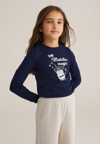 Tengerészeti kék hosszú ujjú póló "Matcha mágia" grafikával és italábrával. Puha anyag, egyszerű szabás, kerek nyakkivágás. Világos nadrággal párosítva.