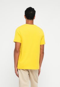 Κίτρινο βαμβακερό T-shirt με στρογγυλή λαιμόκοψη, κοντά μανίκια και άνετη εφαρμογή, φανερό από την πίσω πλευρά, συνδυασμένο με ανοιχτό μπεζ παντελόνι.