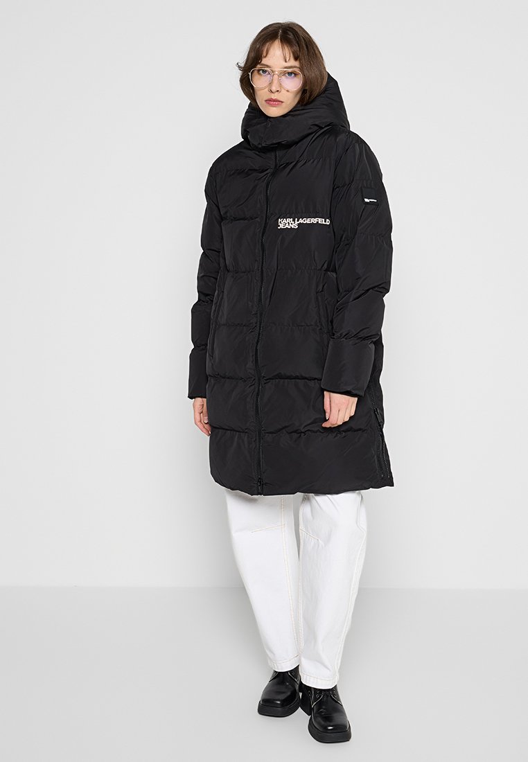 Manteau en duvet noir avec un col haut, fermeture éclair et poches latérales. Présente un logo sur la poitrine et un design matelassé. Porté par-dessus un pantalon blanc.