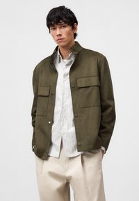 Giacca verde oliva con due tasche frontali, bottoni a pressione e design con colletto, indossata sopra una camicia a righe chiara e pantaloni crema.