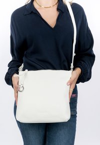 Naine, kes kannab tumesinist pluusi ja teksaseid, hoides valget nahast üle õla rippuvat kotti, millel on hõbedane sõrmusekujuline tõmblukgu tõmblukk.