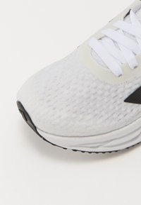 Gros plan sur une chaussure de sport blanche avec une tige en mesh, des lacets blancs et une semelle en caoutchouc noire, mettant en valeur la texture et le détail du logo latéral.