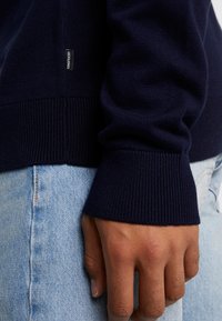 Marineblauer Strickpullover mit gerippten Bündchen, versehen mit einem gewebten Etikett an der Seite. Hand ruht auf leicht blauer Denim-Stoff.