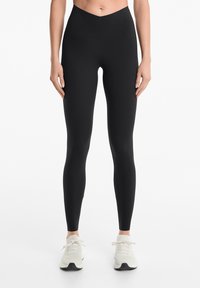 Leggings noirs taille haute en matériau extensible. Présentent une texture lisse et un design ajusté, associés à des chaussures de sport de couleur claire.