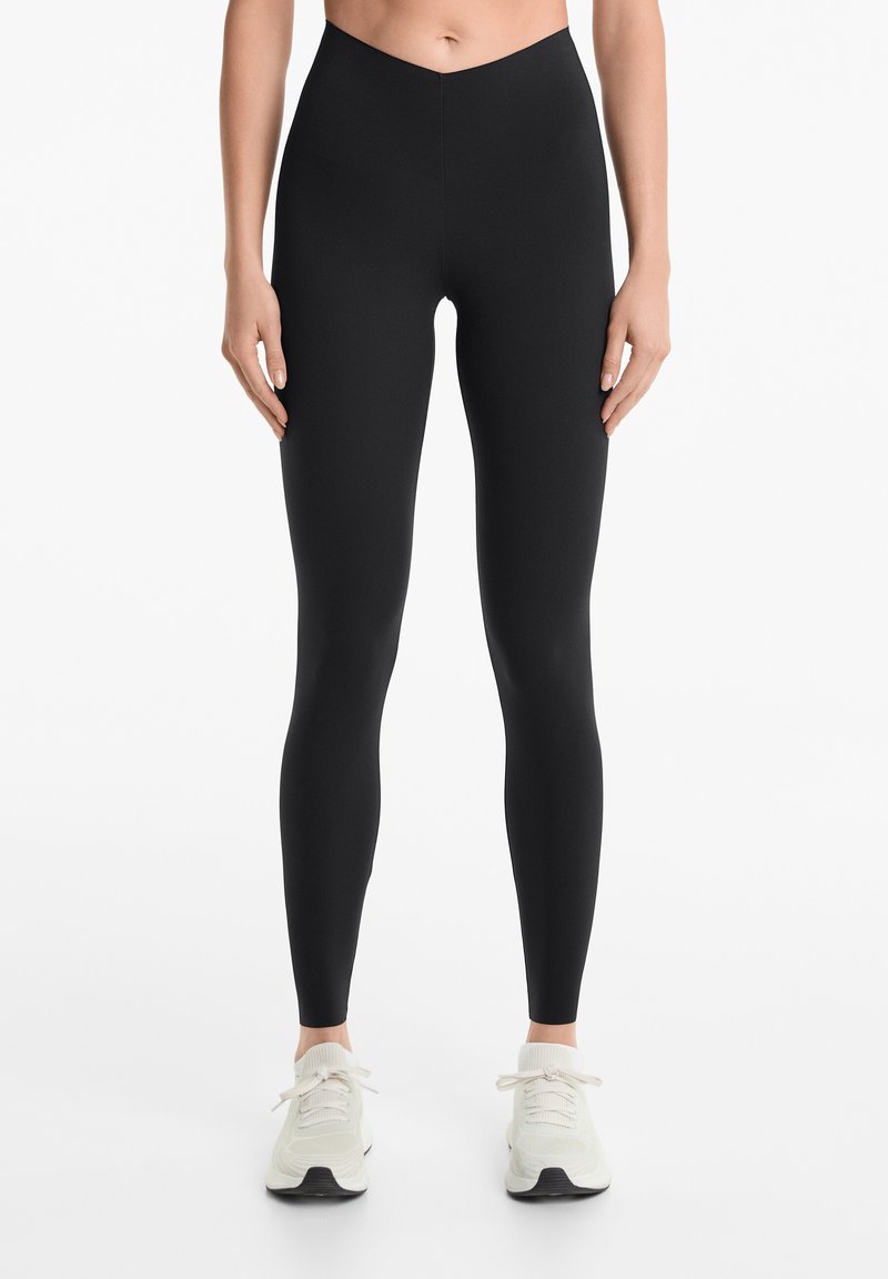 Leggings noirs taille haute en matériau extensible. Présentent une texture lisse et un design ajusté, associés à des chaussures de sport de couleur claire.