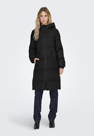 Zwarte pufferjas met capuchon, voorzien van een rits aan de voorkant, twee zijkleppen en een knielange snit, gecombineerd met donkere jeans en boots.