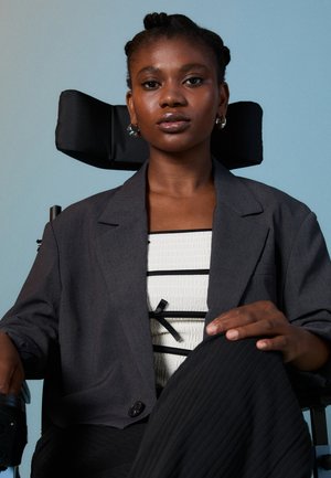 Personne assise dans un fauteuil roulant noir, portant un blazer gris foncé par-dessus un haut blanc texturé avec des rayures noires et un nœud. Pantalon texturé noir.