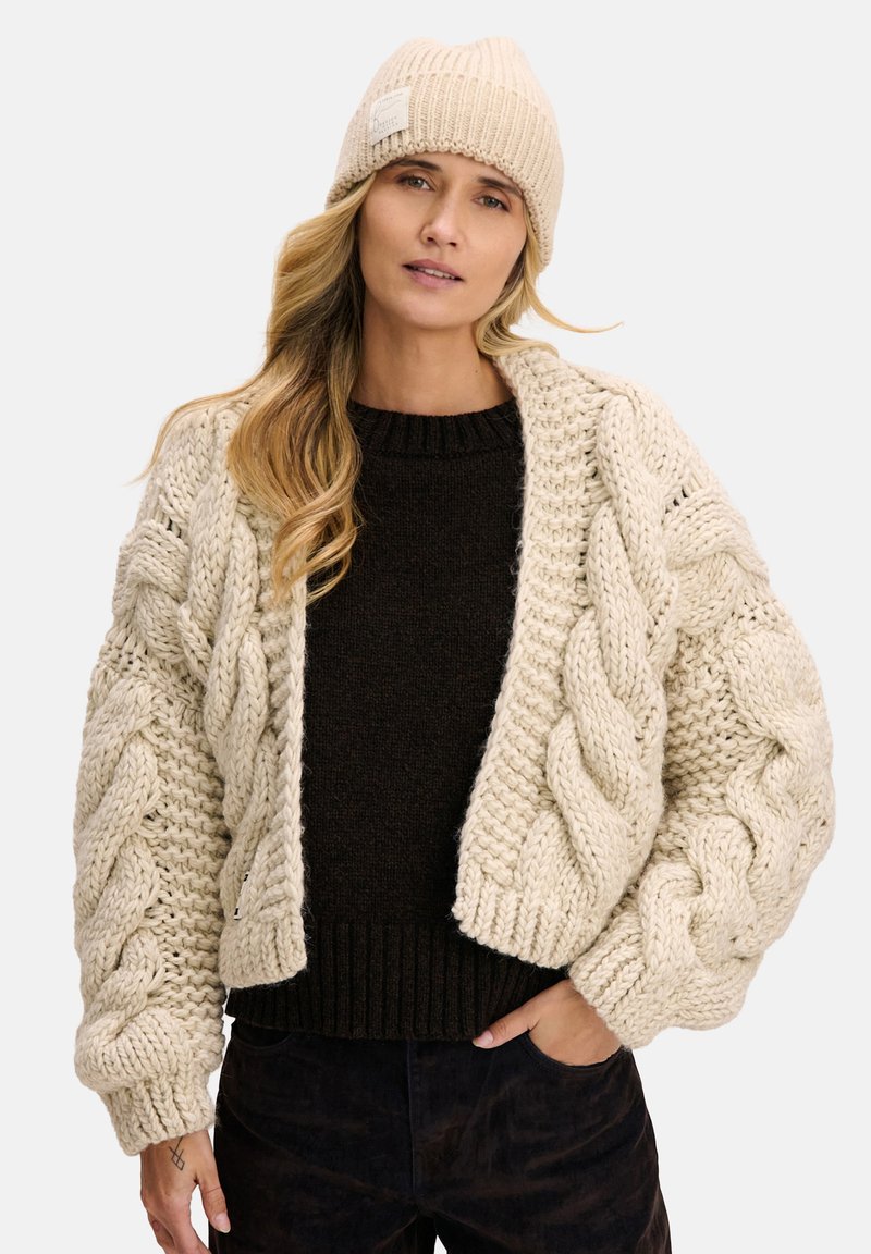 Gruby kremowy kardigan z grubym splotem warkoczowym z dużymi, oversize rękawami, w zestawieniu z ciemnobrązowym, grubym swetrem i jasnobeżową prążkowaną czapką beanie.