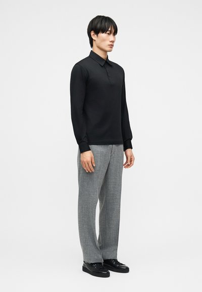 Άνδρας που στέκεται, φορώντας μαύρο πόλο μπλούζα με μακριά μανίκια, γκρι καρό παντελόνι και μαύρα αθλητικά παπούτσια σε λευκό φόντο.
