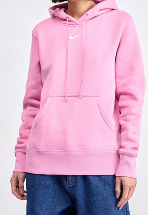 Osoba w różowej bluzie Nike z kapturem, przednią kieszenią i sznurkami, ubrana w luźne niebieskie dżinsy, na jednolitym tle.