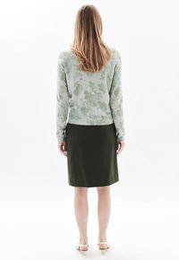 Lichtgroene gebloemde gebreide trui met lange mouwen en geribbelde zoom, gecombineerd met een donkergroene rok tot op de knie. Eenvoudige schoenen met blokhak.