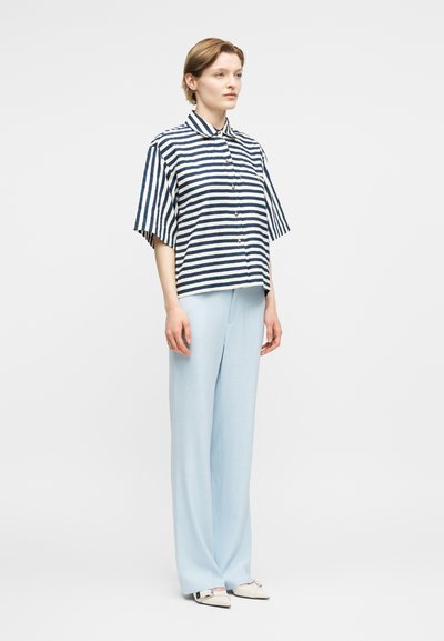 Γυναίκα που φοράει μπλε σκούρο και άσπρο ριγέ πουκάμισο με κοντό μανίκι και κουμπιά, ανοιχτόχρωμο γαλάζιο παντελόνι με φαρδιά μπατζάκια και λευκά μυτερά παπούτσια, στέκεται μπροστά σε λευκό φόντο.