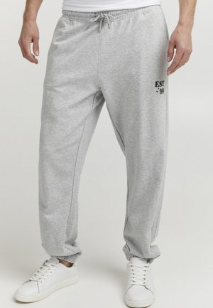 Grijze sportbroek met elastische tailleband en boorden, voorzien van een logo-opdruk "EST '99" in het zwart. Het materiaal lijkt zacht en gestructureerd.