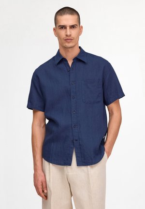 Mann trägt ein marineblaues kurzärmeliges Hemd mit Brusttasche und beiger Hose, steht vor neutralem Hintergrund.