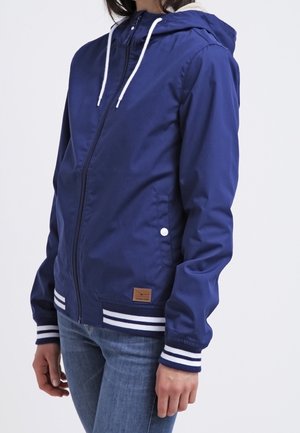 Personne portant une veste à capuche bleu marine avec des cordons blancs, des poignets et un ourlet rayés, ainsi qu'un jean bleu, sur un fond uni.