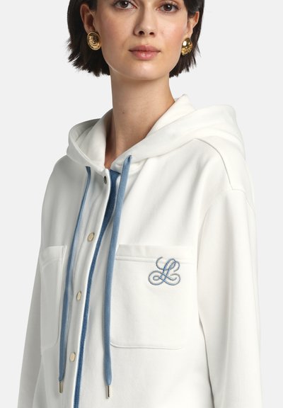 Femeie purtând o jachetă albă cu glugă, cu șnururi albastre și broderie "LL" pe buzunarul de la piept, asortată cu cercei rotunzi aurii.