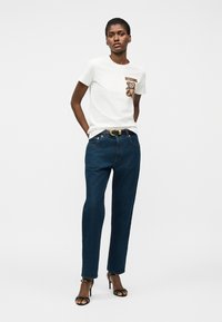 Mujer de pie con camiseta blanca de Moschino con gráfico de oso de peluche, jeans azul oscuro con cinturón y sandalias negras de tacón alto, sobre fondo liso.